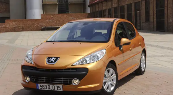 Pomarańczowy Peugeot 207 SW 1.4 benzyna zaparkowany na brukowanym placu przed ceglanym budynkiem.