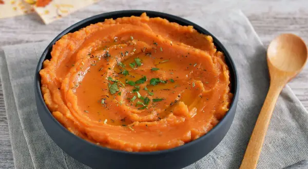 Un tazón de puré de boniato gourmet, cremoso y adornado con aceite y hierbas. Una cuchara de madera descansa a su lado.