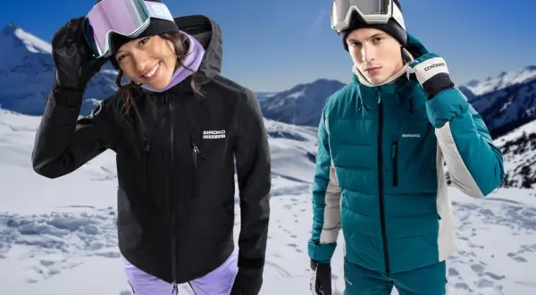 Pareja sonriente en la nieve, ella con chaqueta hardshell negra, él con chaqueta acolchada verde.