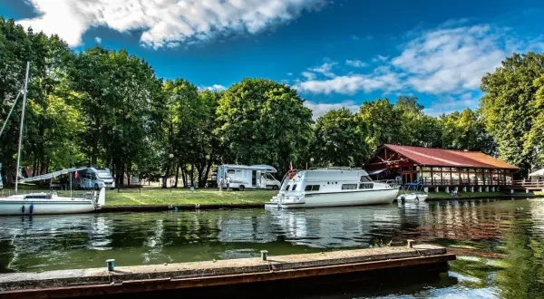 Malownicze pojezierza w Polsce: kampery i łodzie cumują przy brzegu, w tle drewniana restauracja i bujna zieleń.