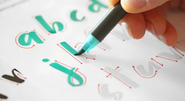 Mano practicando lettering con plantillas imprimibles gratis. Letras "a, b, c, j, i, t, u" en verde y rojo.