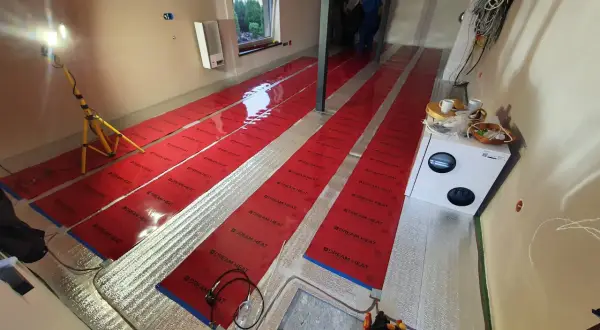 Instalacja folii grzewczej. Widać długie pasy czerwonej folii na podłodze, przygotowane do montażu. Wady tej metody mogą obejmować nierównomierne rozprowadzanie ciepła.