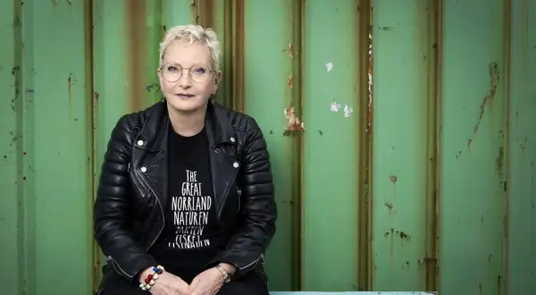 Ola Liljedahl, med kort blont hår och glasögon, sitter framför en sliten grön vägg. Hon bär en svart läderjacka och en t-shirt med texten "THE GREAT NORRLAND NATUREN".