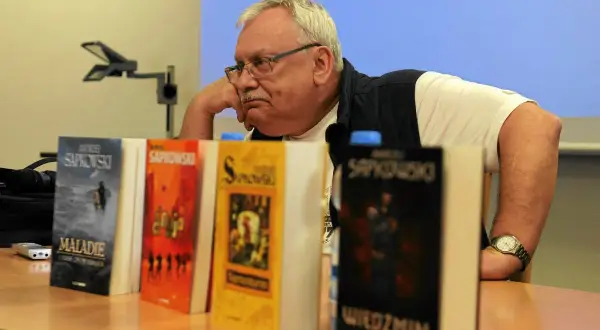 Mężczyzna zamyślony nad stosem książek, wśród nich "Wiedźmin" i inne tytuły Sapkowskiego, idealne dla fanów szukających książek podobnych do Wiedźmina.