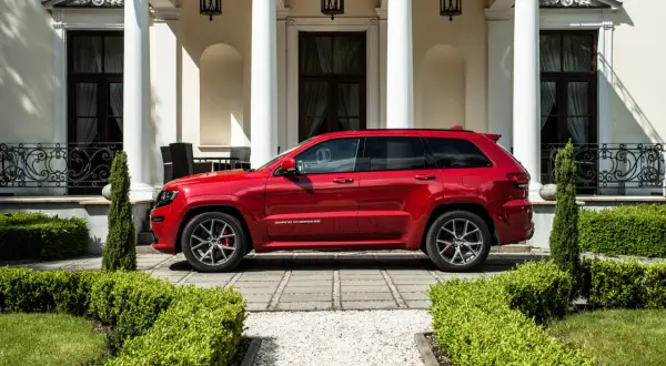 Czerwony Jeep Grand Cherokee SRT zaparkowany przed elegancką posiadłością z białymi kolumnami i zadbanym ogrodem.