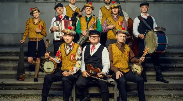 Zespół muzyczny w strojach ludowych z kapeluszami w kratę, grający na instrumentach: banjo, skrzypcach, akordeonie, saksofonie, kontrabasie i bębnie.