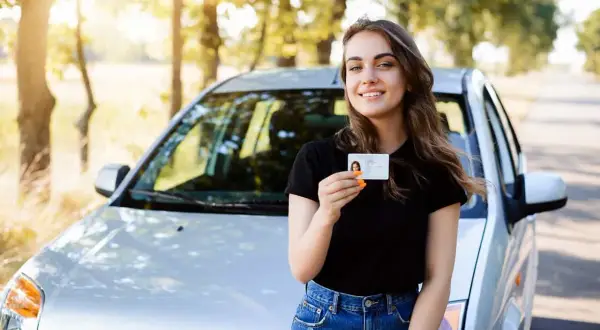 Szczęśliwa młoda kobieta z prawem jazdy od 16 lat stoi obok samochodu na tle drzew.