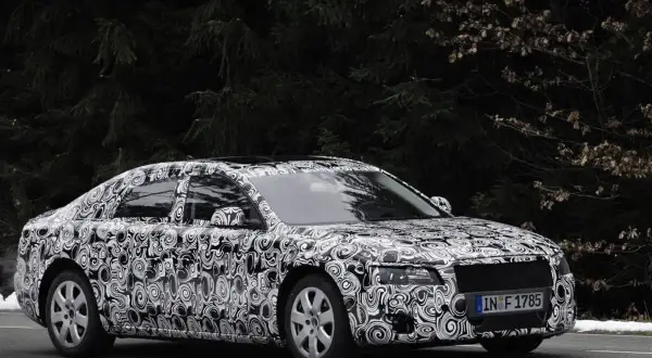 Audi Limousine mit Tarnfolie fährt auf schneebedeckter Straße vor dunklem Wald.