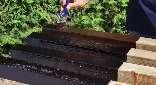 Mano che applica vernice scura su listelli di legno grezzo, un passo essenziale per imparare come impermeabilizzare il legno e proteggerlo.