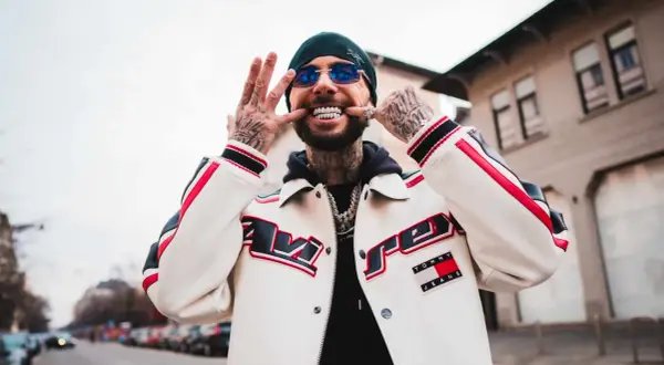 Malik Montana z uśmiechem i błyszczącymi zębami, w kurtce Tommy Jeans. Jego wzrost jest imponujący, a tatuaże dodają mu charakteru.