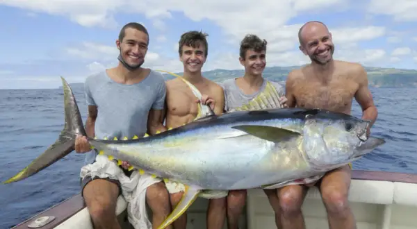 Vier Männer präsentieren stolz einen riesigen Thunfisch auf einem Boot vor den Azoren. Ein erfolgreicher Tag beim **Azoren Angeln**!