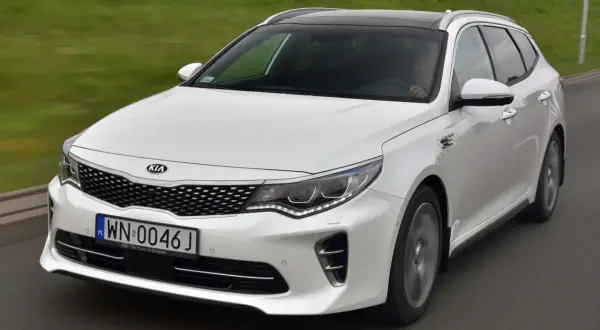 Biała Kia Optima w wersji kombi na drodze. Wiele pozytywnych kia optima opinie potwierdza jej wszechstronność.