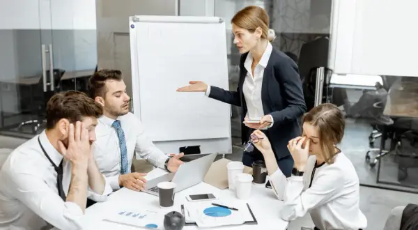 En kvinna gestikulerar mot en whiteboard medan kollegor ser utmattade ut. Känslan av manipulativt beteende hänger i luften under mötet.