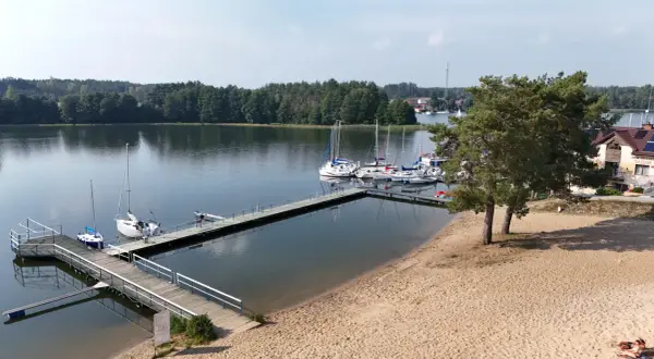 Wdzydze Kiszewskie plaża z pomostami i żaglówkami. Piasek, woda i zielone drzewa tworzą idylliczny krajobraz.