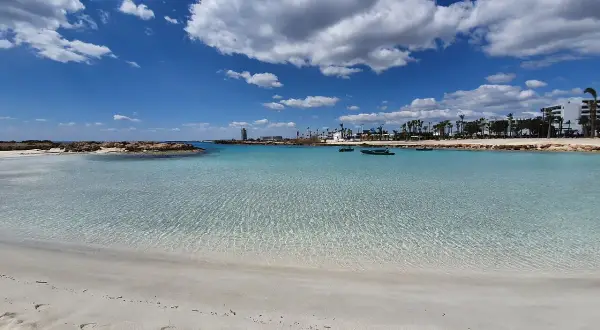 Kristallklart vatten och vit sand vid Nissi Beach, Cypern. Små båtar guppar i det turkosa vattnet under en blå himmel med vita moln.