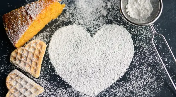 A slice of cake, heart-shaped cookies, and a heart shape made of powdered sugar on a dark surface.