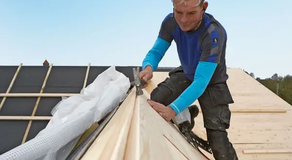 A roofer wearing safety glasses hammers a wooden plank onto a roof under construction.