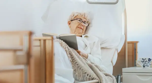 Ältere Dame liest im Bett, eine Auszeit in der Kurzzeitpflege ohne Pflegegrad. Gemütlich eingekuschelt mit einem Buch.