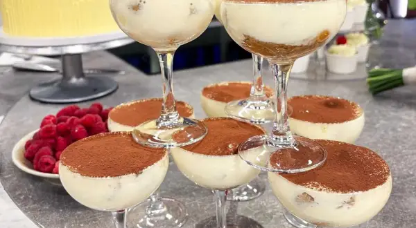 Flera glas med krämig tiramisu, pudrad med kakao. Hallon och en gul tårta i bakgrunden.