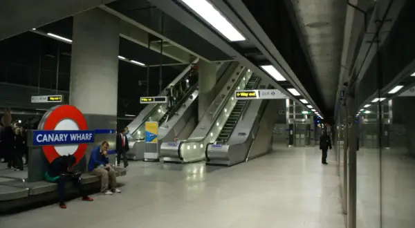 Stacja Canary Wharf w **metro londyn** z charakterystycznym znakiem, ruchomymi schodami i ludźmi czekającymi na pociąg.