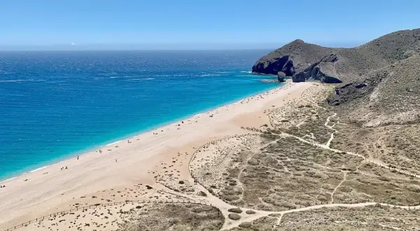 Una cala de arena dorada se extiende junto a un mar turquesa. Senderos serpentean por las colinas áridas que rodean la playa.