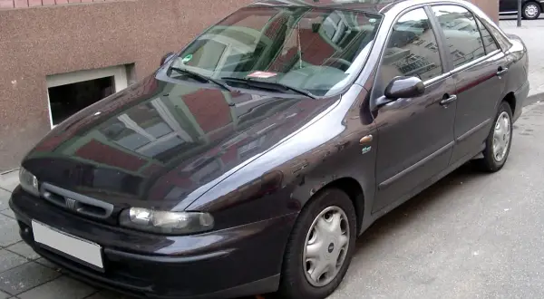 Ciemny Fiat Marea stoi zaparkowany przy ceglanym budynku.