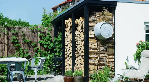Vedbod med snyggt staplad ved. En utomhusplats med bord, stolar och en grill.