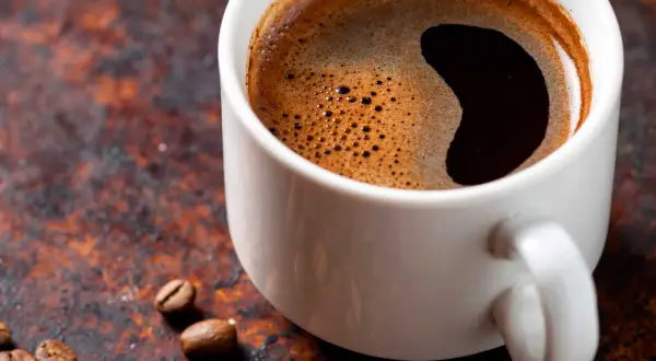 En vit kopp med mörkt kaffe och skum. Kaffebönor ligger utspridda. Undrar du hur mycket koffein i en kopp kaffe?