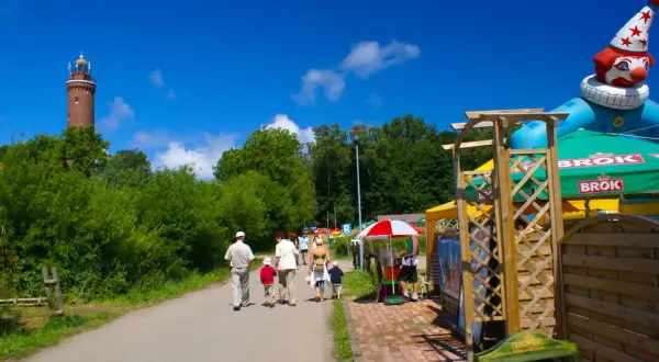 Rodzina spaceruje ścieżką w Ustroniu Morskim w kierunku latarni. W tle wesoły klaun i zielone parasole.