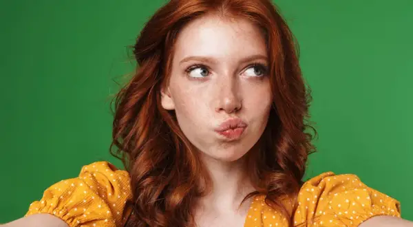 A young woman with fiery red hair, often symbolizing passion and uniqueness, puckers her lips against a vibrant green backdrop.