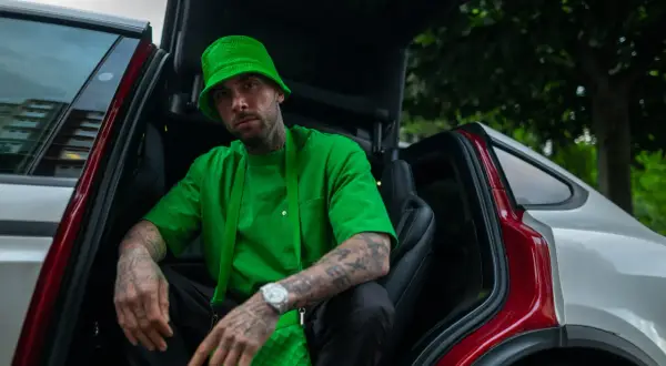 Malik Montana w zielonym kapeluszu i koszulce, siedzi w samochodzie z otwartymi drzwiami. Wygląda jak adwokat diabła, gotowy na nowe wyzwania.