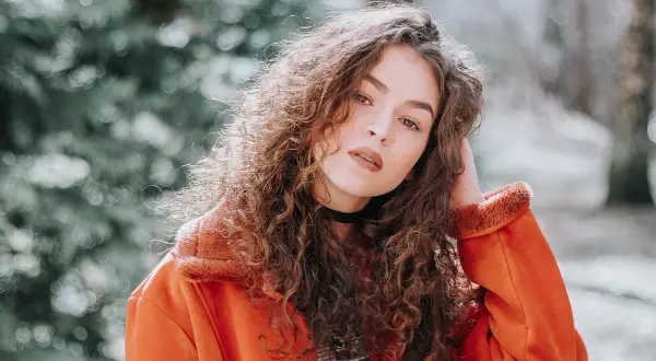 Joven con melenas rizadas y un abrigo naranja, posando con confianza.