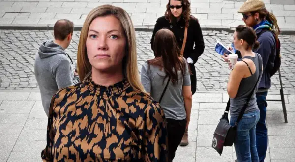En kvinna med en mönstrad blus tittar framåt. I bakgrunden syns flera personer, en del verkar diskutera, vilket kan symbolisera ansvarstagande på jobbet.