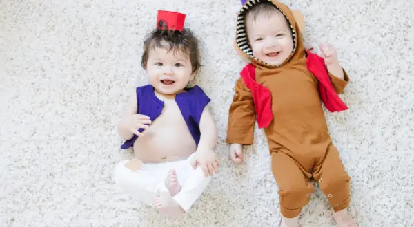 Dwa uśmiechnięte niemowlaki w strojach inspirowanych Alladynem leżą na białym dywanie. Jeden w fioletowej kamizelce, drugi w brązowym kombinezonie.