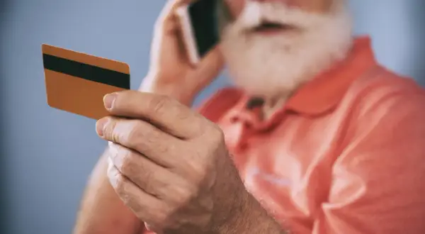 Starszy mężczyzna z brodą trzyma kartę i rozmawia przez telefon, zastanawiając się, dlaczego bank zablokował kartę.