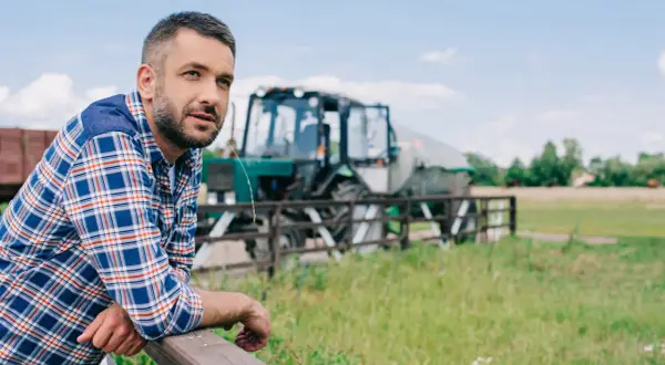 Rolnik w kratę oparty o płot, w tle traktor. Wsparcie dla małych gospodarstw to klucz do rozwoju wsi.