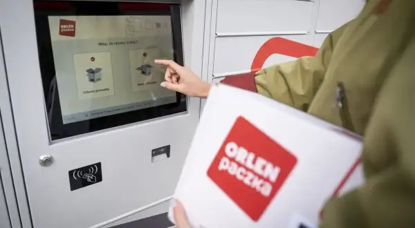 Kobieta z paczką ORLEN PACZKA dotyka ekranu automatu, wybierając opcję nadania przesyłki.
