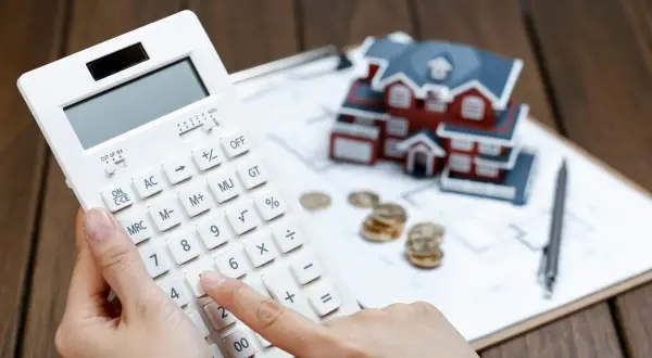 Calculadora en mano, se evalúa cuánto se deduce por alquiler de vivienda, con una maqueta de casa, monedas y planos.