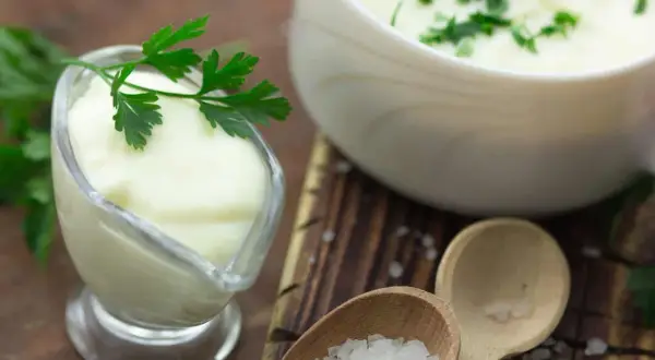 Cremige Bechamel-Sauce in einer Sauciere, garniert mit Petersilie. Daneben eine Schüssel mit der Sauce und grobes Salz auf Holzlöffeln.