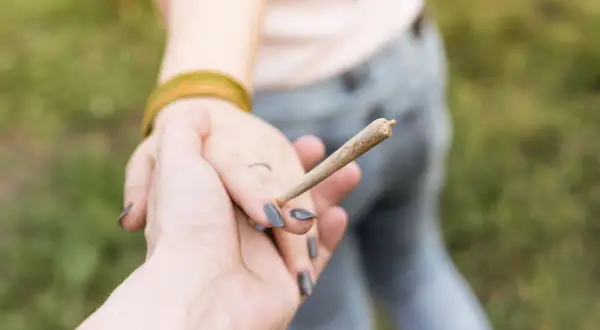 Dłoń trzymająca skręta, symbol naturalnych narkotyków, w tle druga osoba.