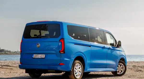Błękitny VW Transporter T7 na piaszczystej plaży, z widokiem na morze.