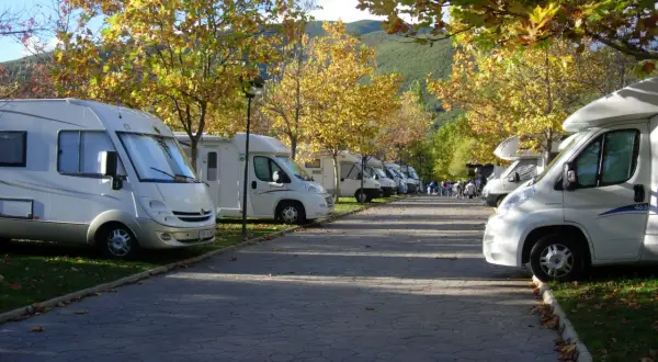 Un tranquilo caravan parking bajo árboles otoñales. Varias autocaravanas blancas aparcadas en fila, con hojas doradas cayendo.