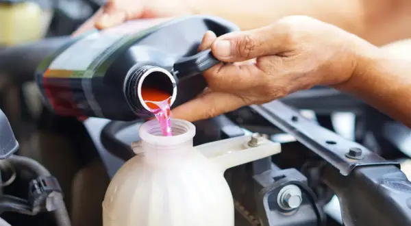 Eine Hand gießt rosa Kühlflüssigkeit aus einem schwarzen Behälter in einen weißen Ausgleichsbehälter eines Autos.