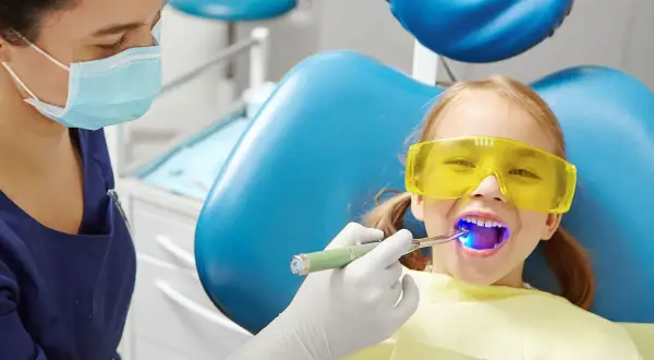 Dziecko w okularach ochronnych uśmiecha się, gdy dentystka utwardza lakowanie zębów. Czy lakowanie zębów jest szkodliwe? Nie, to bezpieczna profilaktyka.