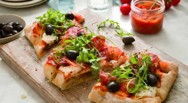 Pinsa med salami, ruccola och oliver på en träskärbräda. Bredvid står en burk tomatsås och en skål med oliver.