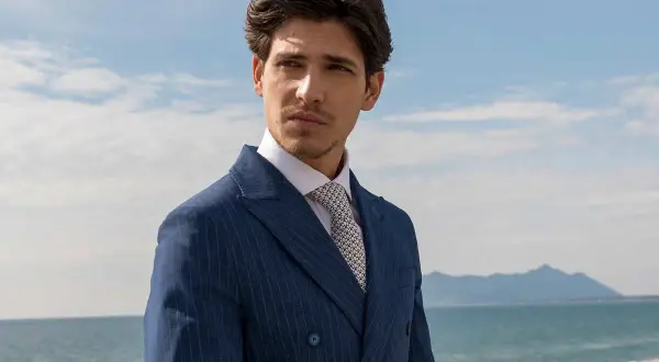 Uomo con abito blu gessato e camicia bianca, indossa una cravatta con un elegante nodo cravatta simmetrico. Sguardo pensieroso verso l'orizzonte marino.