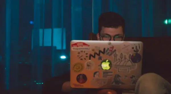 Chłopak z okularami szuka angielskich nicków do gier na laptopie ozdobionym naklejkami.