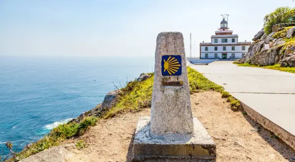 Kamienna wskazówka z symbolem muszli i napisem "0.000 KM" na trasie Camino Portugues. W tle biała latarnia morska i błękitne morze.