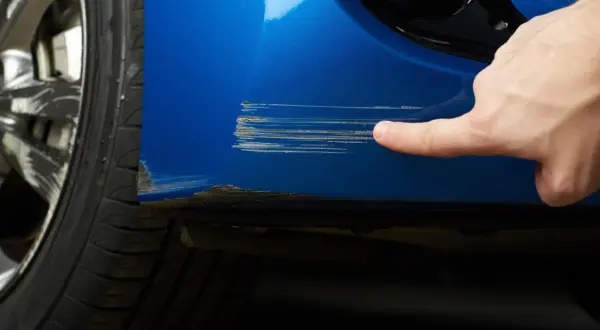 Dito si vede come riparare graffi auto. Un dito indica profondi graffi su un'auto blu vicino alla ruota.