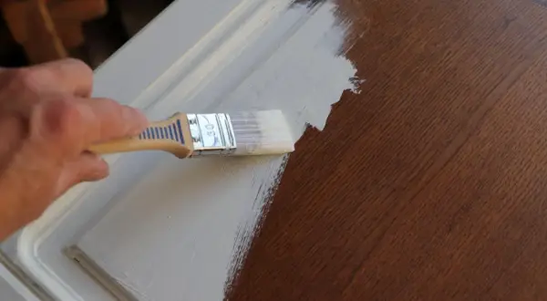Main d'une personne appliquant une couche de peinture claire sur un meuble en bois verni.
